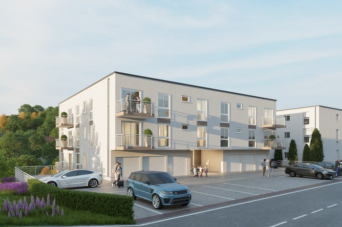 Modernes Mehrfamilienhaus mit drei Stockwerken, Balkonen und gepflastertem Parkplatz mit Autos.