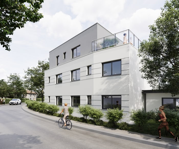 Modernes, vierstöckiges Wohngebäude mit Flachdach, umgeben von Bäumen und Büschen, an einer Straße.
